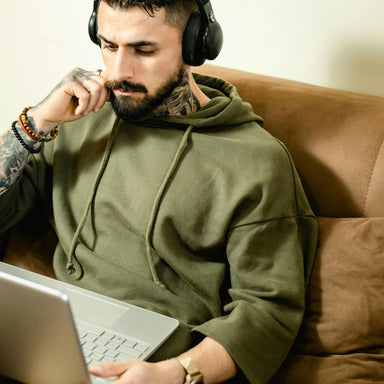A person with tattoos is seated on a couch, wearing headphones and using a laptop in their lap.